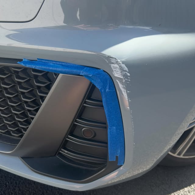Close view of a grey front bumper corner with masking tape around a scuffed edge.